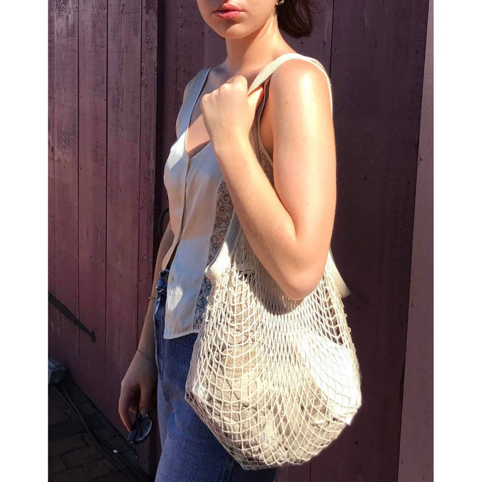 A woman wearing a white sleeveless top and blue jeans carries a large cream-colored open-knit mesh tote bag against a dark wooden plank wall.