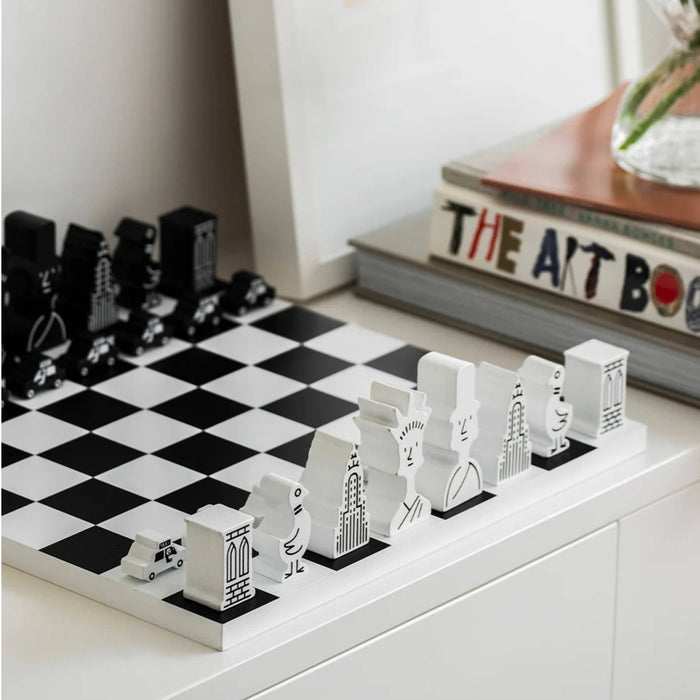 A black-and-white chess set with stylized architectural and vehicle-shaped pieces arranged on a white tabletop beside stacked books.
