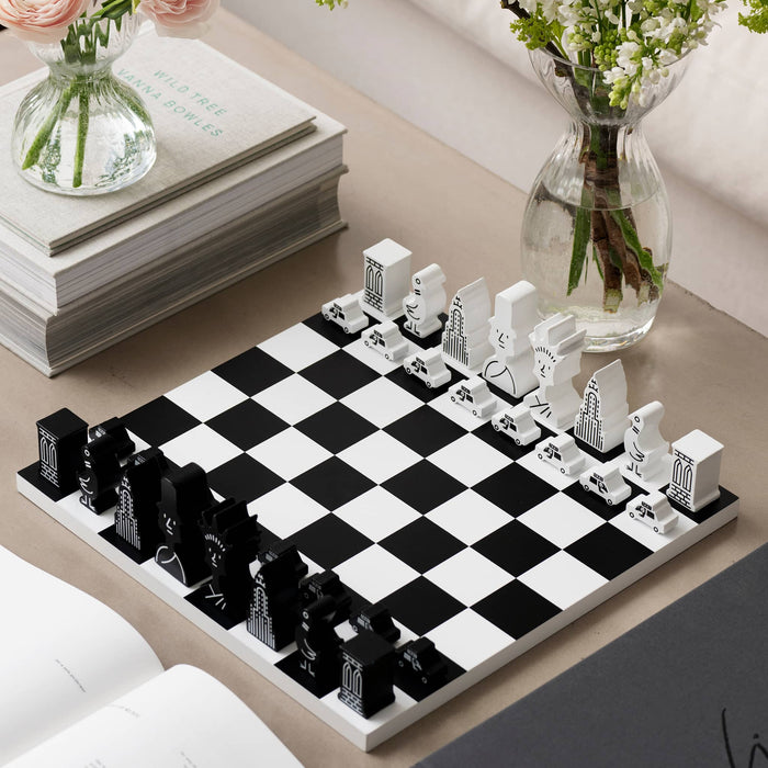 A black and white chess set arranged on a tabletop, with decorative books, flowers in glass vases, and an open book in the background. The chess pieces are designed as New York City icons in black and white.