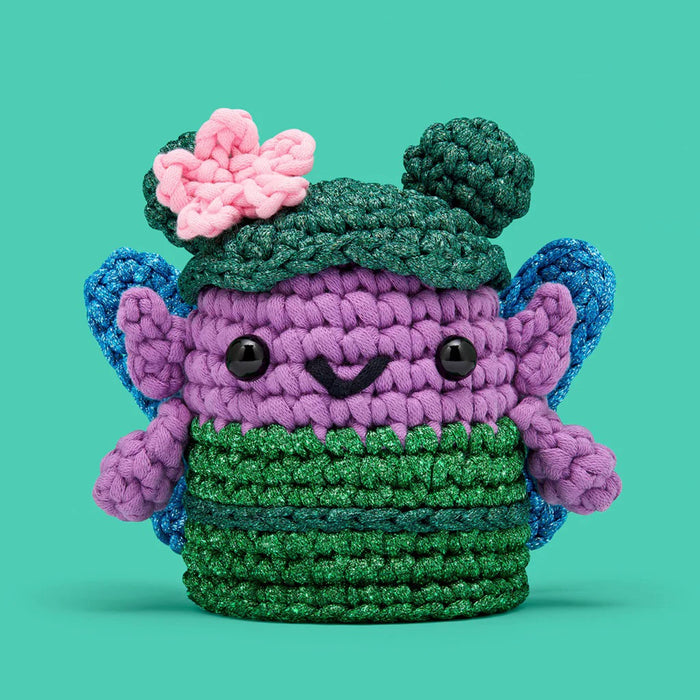 Crocheted fairy character with a green hat and pink flower on a teal green background.