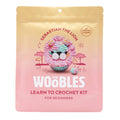 Front of pink and gold ombre pouch packaging for The Woobles Pastel Sebastian the Lion beginner crochet kit, on a white background.