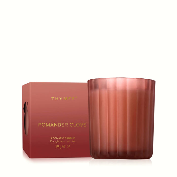 A Thymus Pomander Clove scented candle in a ribbed, translucent soft red glass jar next to its matching red packaging box with gold lettering, set against a clean white background.