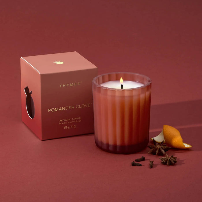 A lit Thymés Pomander Clove scented candle in a ribbed glass jar sits next to its red ombre packaging box on a matching dusty red background, styled with star anise, cloves, and a citrus slice.