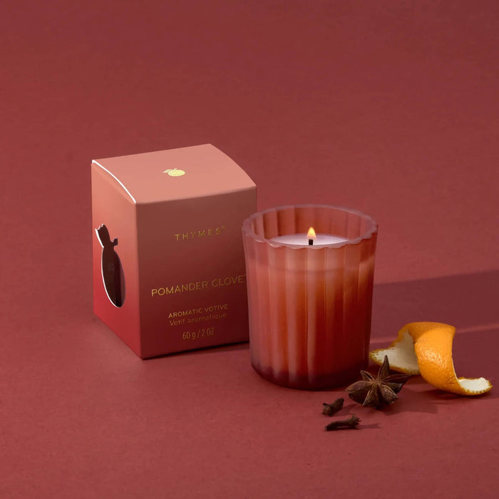 A lit soft red glass votive candle with a small flame stands beside its matching red ombre box on a dusty rose background, with orange peel and star anise props nearby. The box reads 'THYMES' and 'POMANDER CLOVE'.