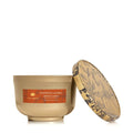 A Thymes 'Pumpkin Laurel' scented candle in a matte gold jar with its lid leaning against it. The lid features a decorative pattern of pumpkins and autumn leaves in black and beige.