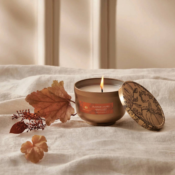 A lit candle in a metallic gold tin sits on draped cream fabric with autumn leaves and small dried botanicals beside it. The lid rests beside the vessel, and the label reads Pumpkin Laurel.