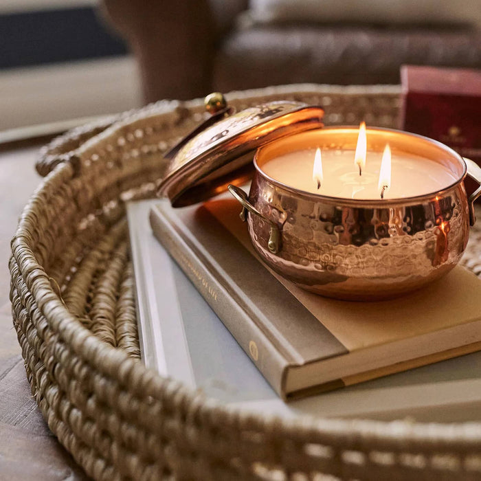Simmered Cider scented candle in copper pot, lid off and 3 wicks lit, sitting on a book in a woven basket.
