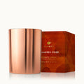 Thymes Simmered Cedar scented candle in metallic rose gold vessel beside illustrated red gift box with gold foil lettering.