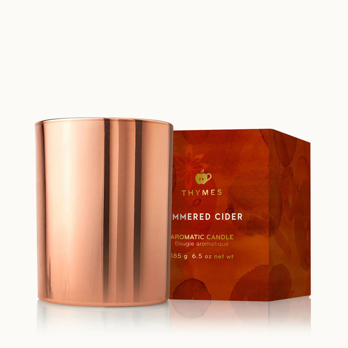 Thymes Simmered Cedar scented candle in metallic rose gold vessel beside illustrated red gift box with gold foil lettering.