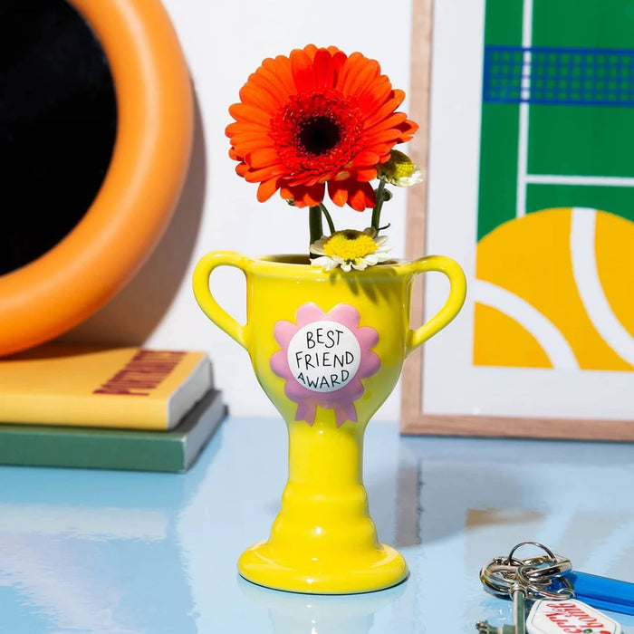 A bright yellow ceramic trophy-shaped vase with two handles, holding an orange gerbera daisy and a small yellow and white flower. A pink ribbon badge on the front reads 'BEST FRIEND AWARD.