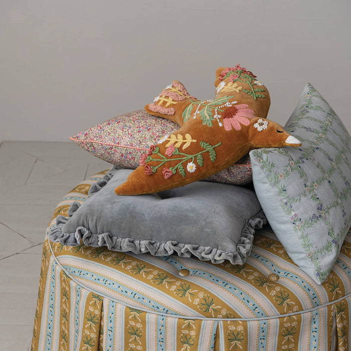 A styled interior scene showing a decorative mustard yellow velvet pillow with floral embroidery resting on a stack of patterned and solid throw pillows atop a striped upholstered bench against a plain light gray wall.