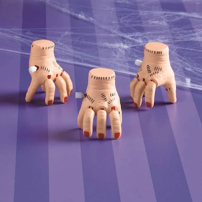 Three severed hand toys with flesh-toned skin, black stitched details, and red fingernails, arranged on a purple striped surface with cobweb graphics.