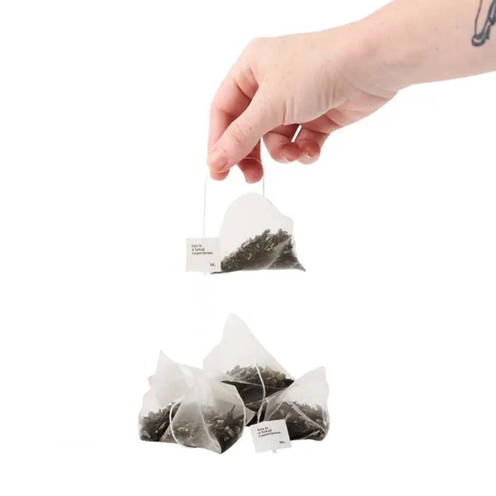 Hand holding a tea bag with black tea leaves against a white background.