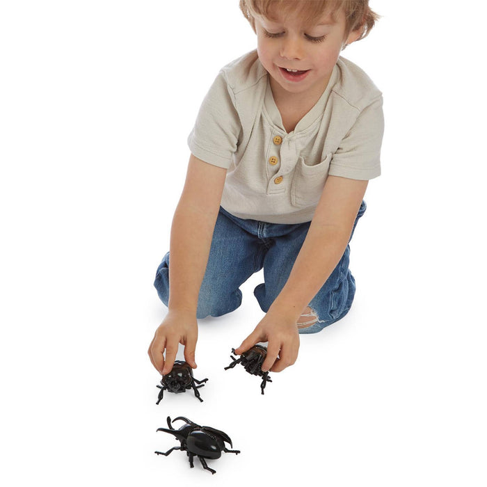 A young child kneeling on a white background, holding and playing with three black plastic beetle toy bugs.