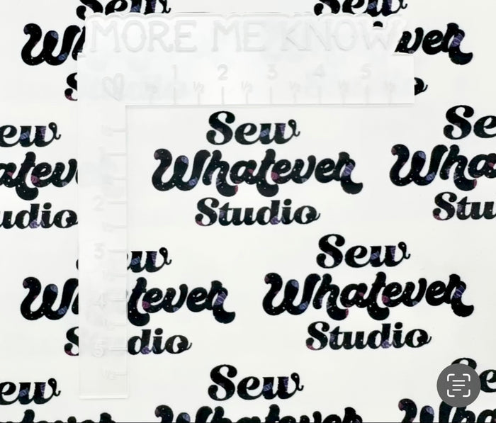 This printed WPCotton features the "Sew Whatever Studio" logo in black with multicolored spools of thread in the wording on a white background. This photo shows a white L shaped ruler or scale.