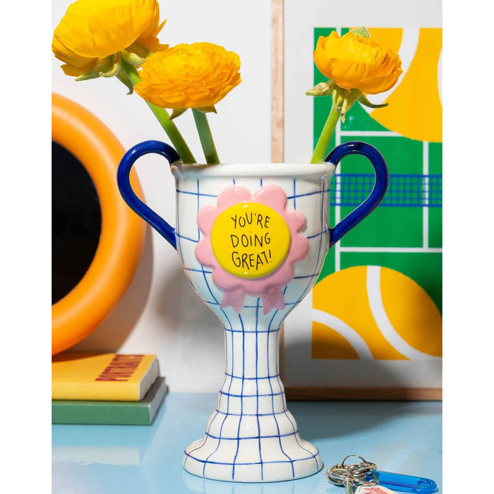 A ceramic trophy-shaped vase with blue handles and a blue grid pattern, holding three yellow flowers. The front has a pink rosette with a yellow center that reads 'YOU'RE DOING GREAT!'.