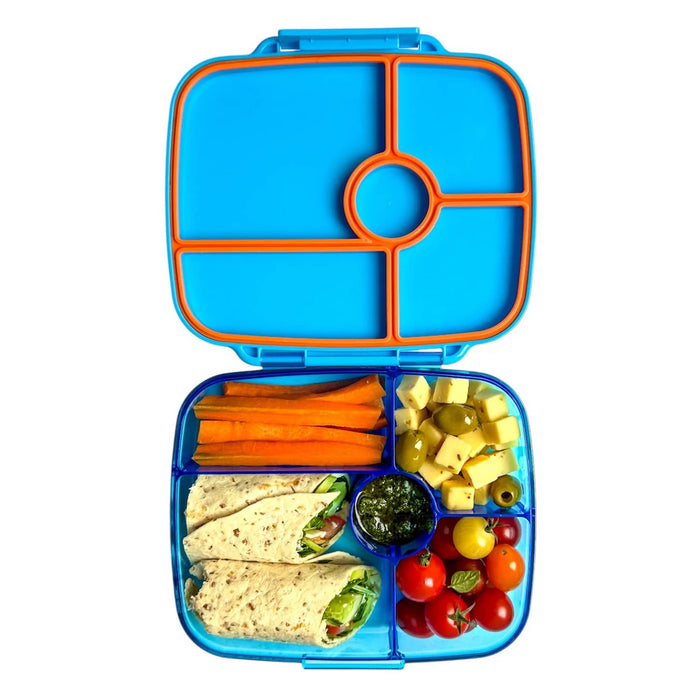 An open blue bento lunch box with orange-trimmed compartments, shown on a white background. The box is filled with a wrap, carrot sticks, cheese cubes, olives, a small green dip, and cherry tomatoes.