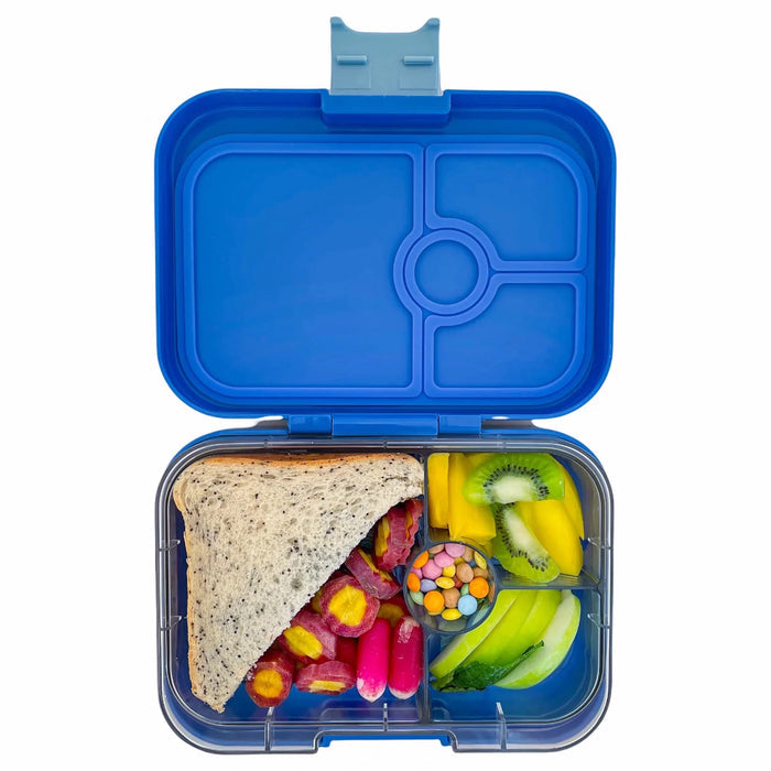 yumbox lunch box in royal blue, lid open showing clear inner tray divided into 4 compartments with a 1/2 sandwich, cut vegetables, chocolate candies, sliced apples, kiwi and mango